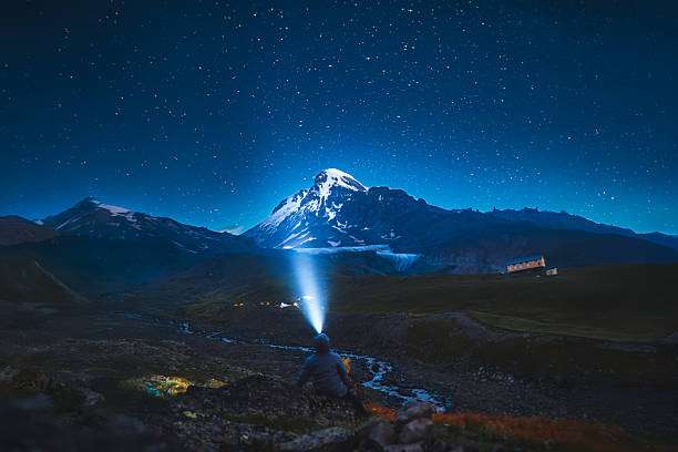 Person uses headlamp under starry night sky with mountains.