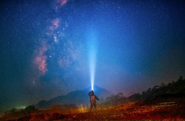 Hiker illuminates starry night sky with a powerful headlamp beam, vast landscape background.