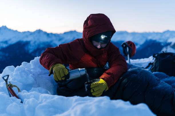 Mountaineer with headlamp pours drink from thermos in snowy mountain landscape.