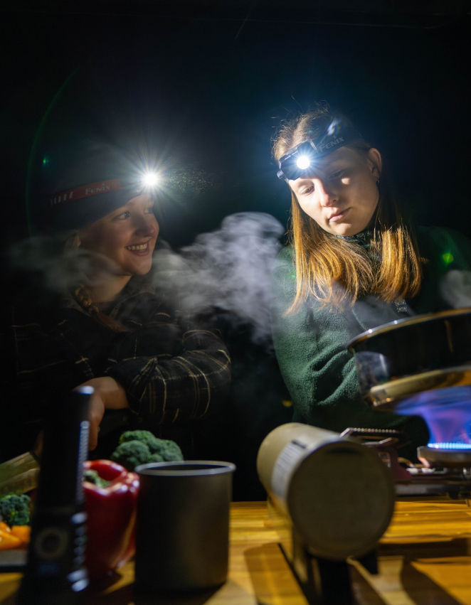 Two women cooking outdoors at night wearing headlamp illumination
