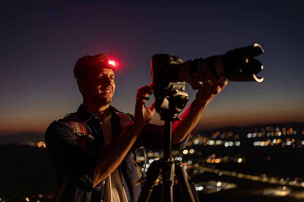 Photographer with a red headlamp adjusts camera on tripod, capturing city lights at night.